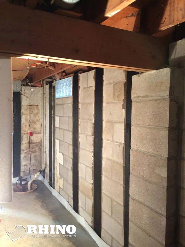 Basement block walls reinforced with vertical black carbon fiber wall supports, with a sump pump in the corner.
